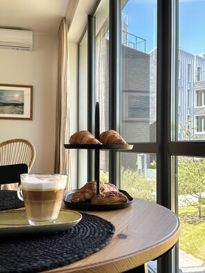 Een tafel met een display van croissants en een koffiekop, een rieten stoel, en een uitzicht op gebouwen door grote ramen.