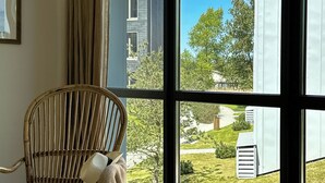 Ein Zimmer mit großem Fenster, einem Korbstuhl und Blick auf ein modernes Gebäude sowie Grünflächen.