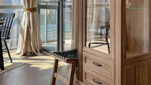 A wooden cabinet with glass doors, a black chair, and a beige curtain in a room with large windows.
