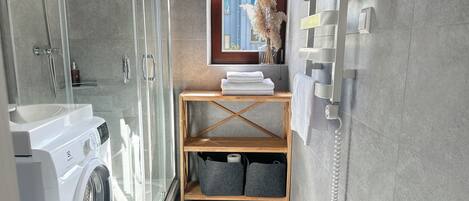 A modern bathroom with a glass shower enclosure, a built-in washing machine, a wooden shelf with towels, and a window with a view of the city.