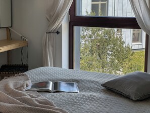 Un dormitorio con una cama, un libro, un escritorio y una ventana con vistas a árboles.