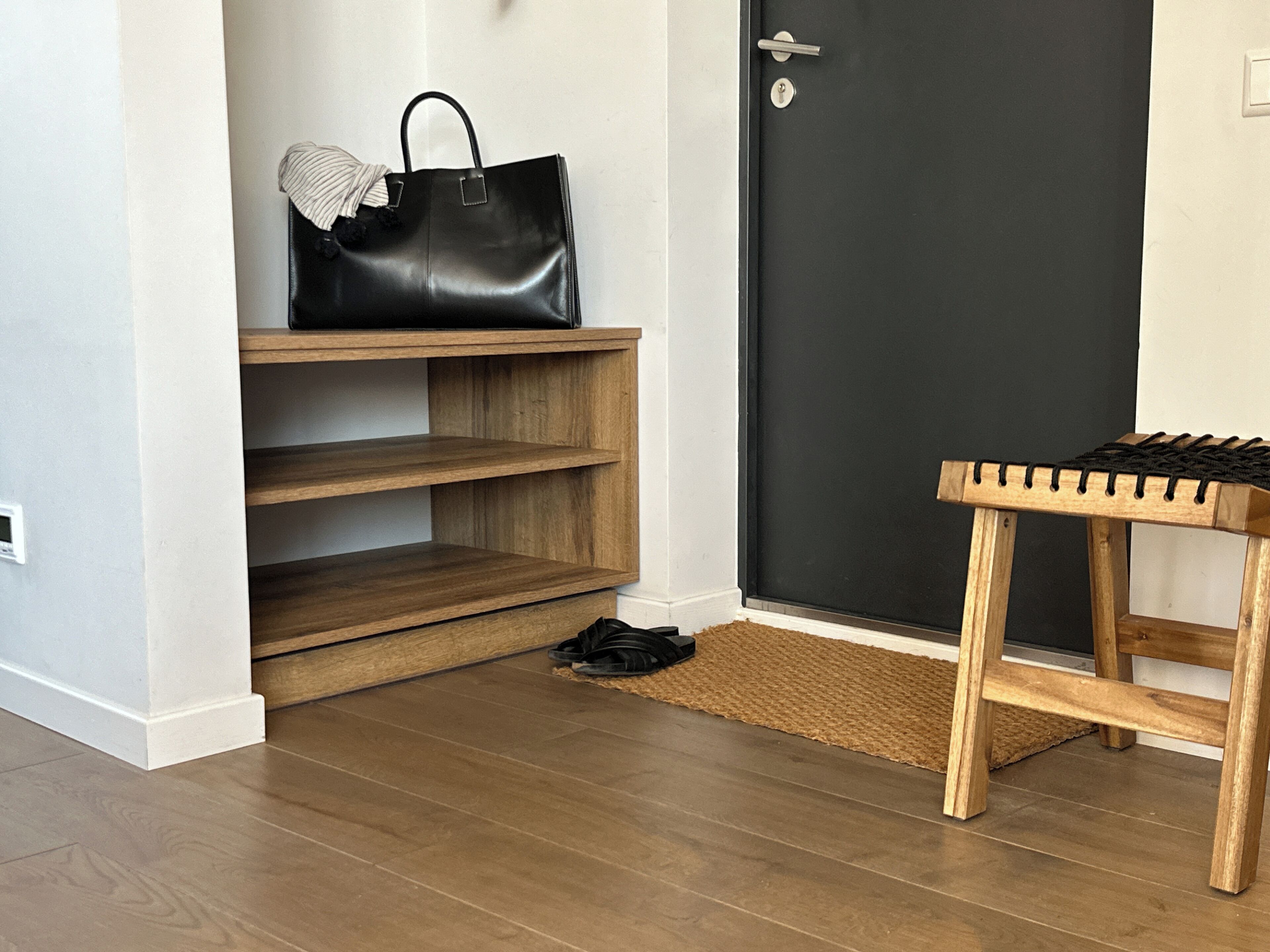 Un couloir avec un banc en bois, un sac à main noir posé sur une étagère et une porte noire.