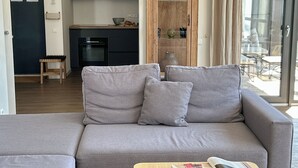 Una sala de estar con un sofá gris, un mueble de madera con puertas de vidrio y una cocina al fondo.