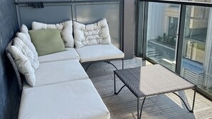 Un balcón con sofá, una mesa pequeña, ventanas amplias y vistas a edificios.