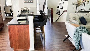 Eine moderne Küche mit Kochinsel, ein Essbereich mit Barhocker, ein Wohnzimmer mit Sofa und einem großen Fenster sowie eine Treppe zu einer weiteren Ebene.