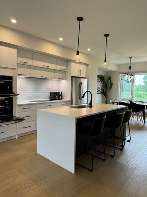 Une cuisine moderne avec des meubles blancs, un îlot central doté de tabourets, un réfrigérateur en acier inoxydable et des luminaires suspendus.