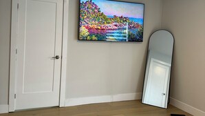 Een slaapkamer met een houten vloer, een bed, een witte deur, een grote spiegel en een kleurrijk schilderij aan de muur.