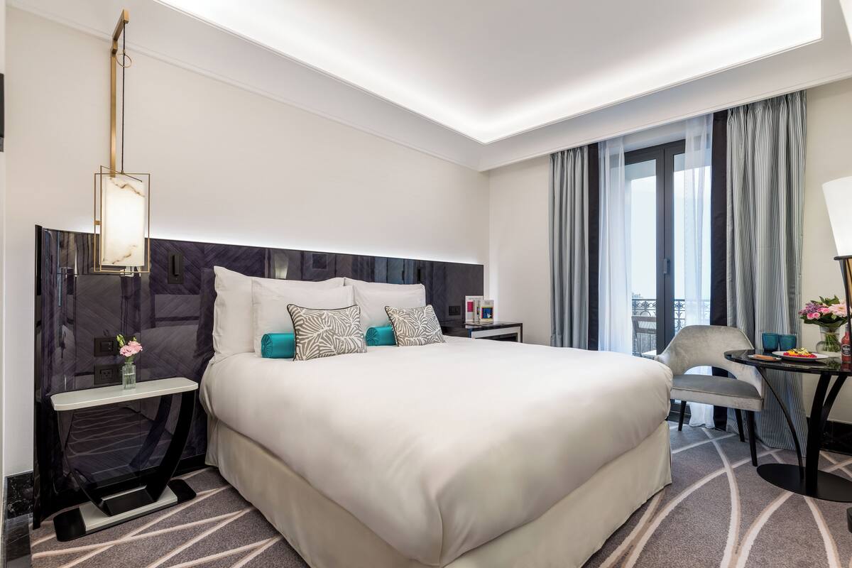 Habitación de hotel moderna con una cama grande, una mesita de noche con un jarrón de flores, una silla y una pequeña mesa con una lámpara y flores.
