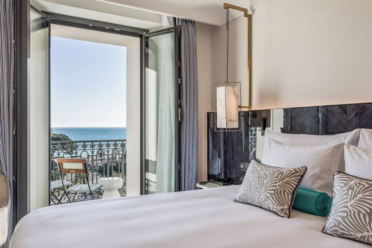Habitación de hotel con una cama grande, vista al océano y balcón con mesa y sillas.