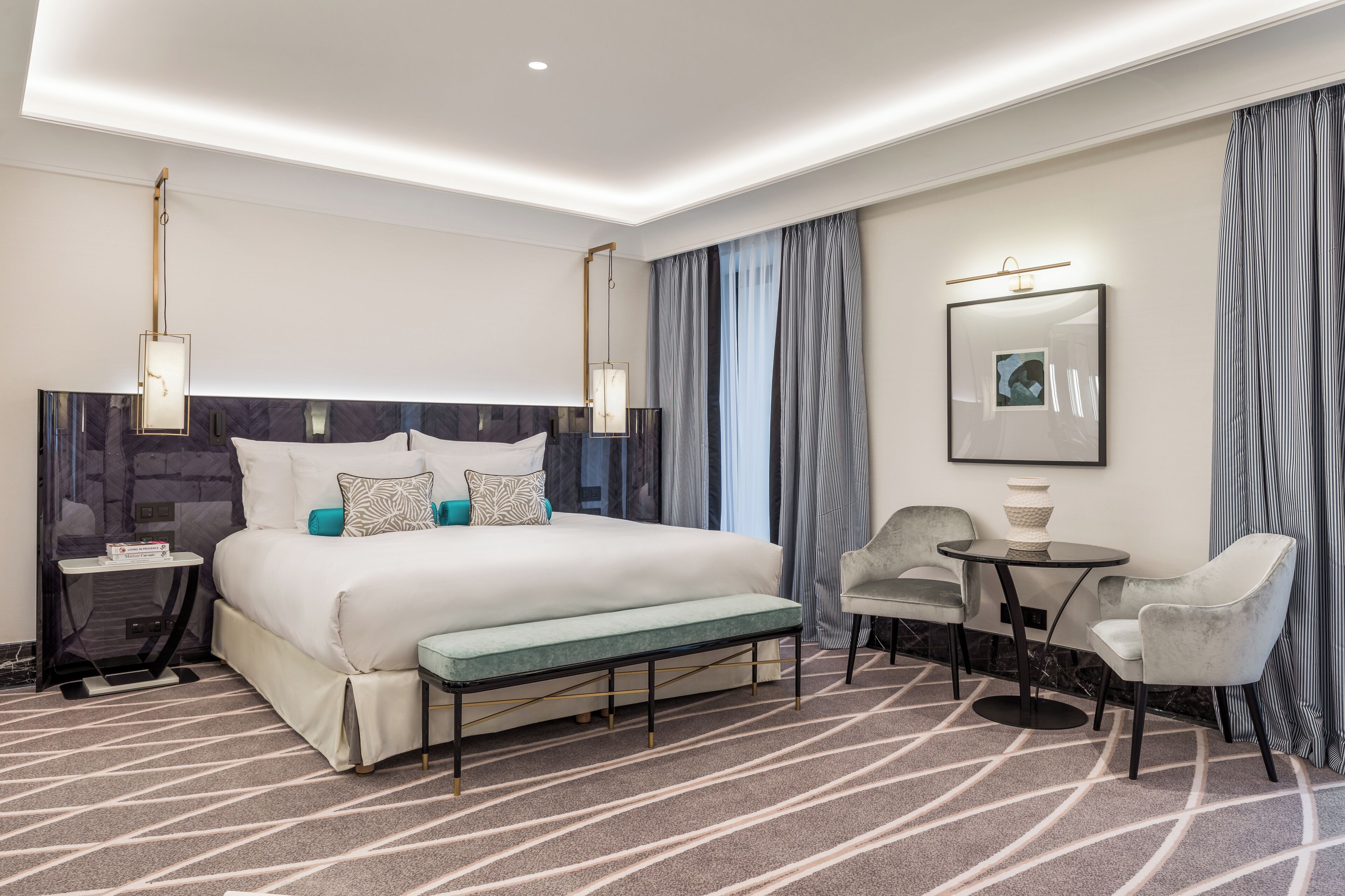 A modern hotel room with a large bed, a bedside table with a lamp, a small round table with chairs, and a large window with curtains.
