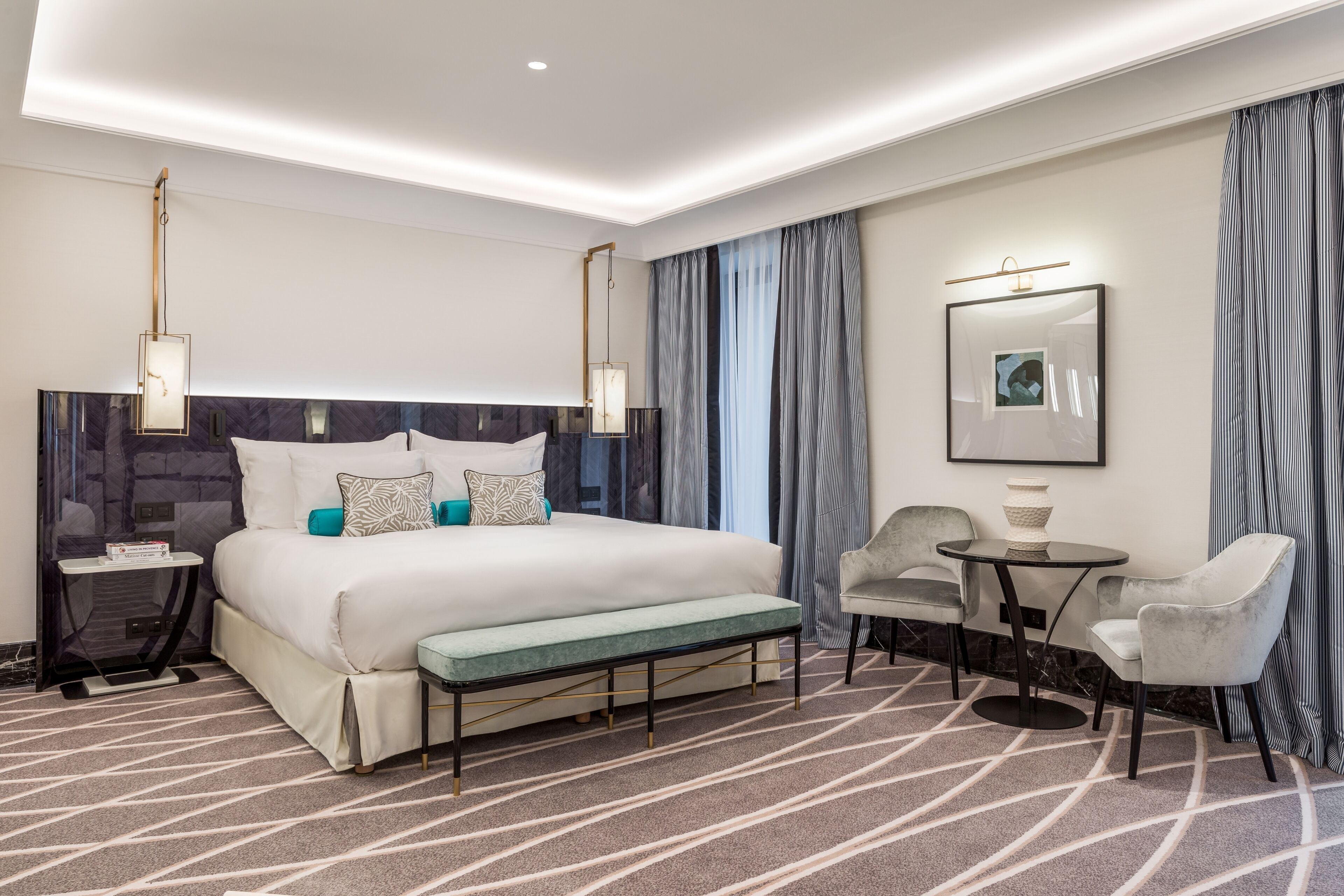 Ein modernes Hotelzimmer mit einem großen Bett, Nachttischen, einer Sitzecke mit Tisch und Stühlen sowie einem großen Fenster mit Vorhängen.