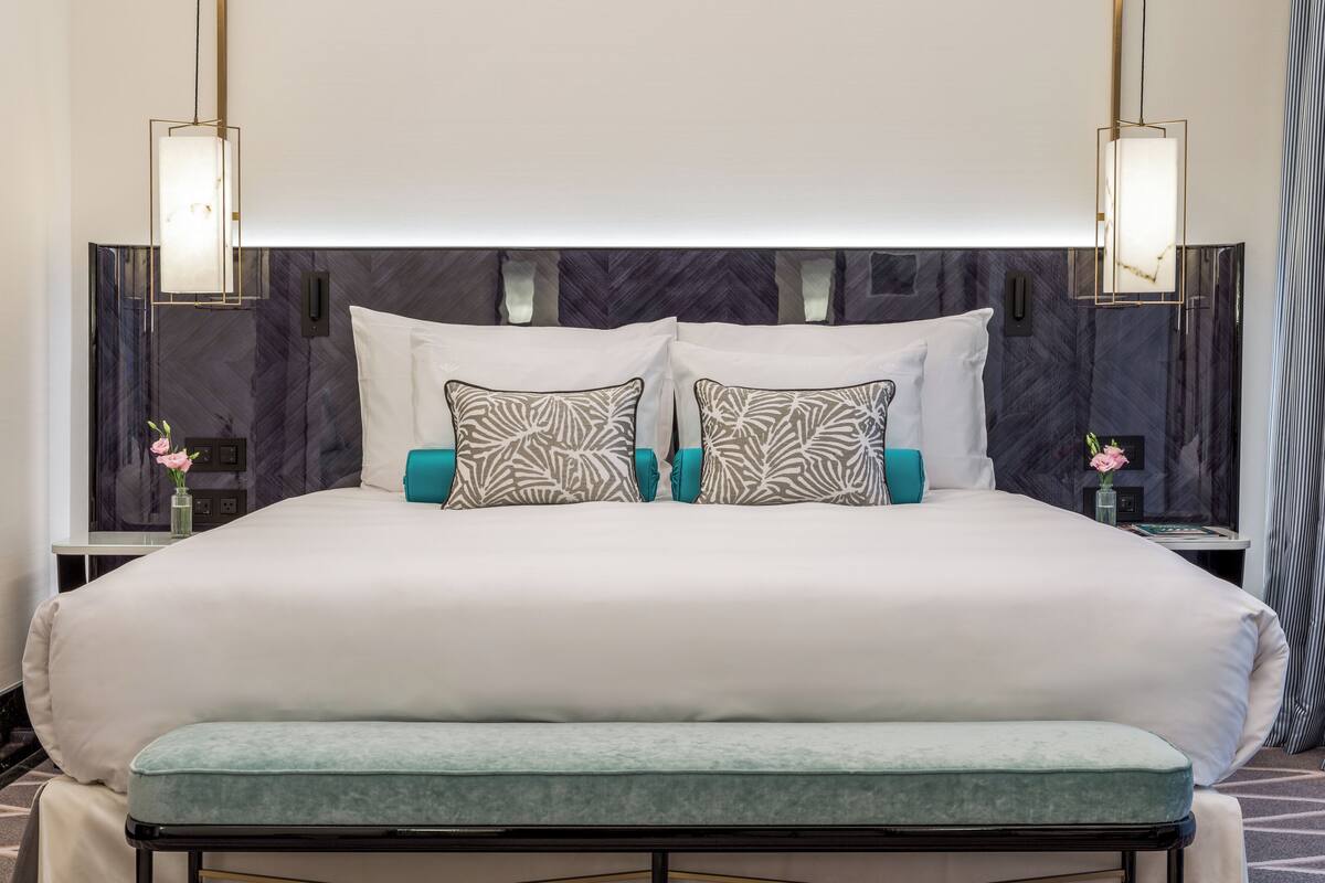 Una cama grande con ropa blanca, almohadas color turquesa y un banco a juego. Hay dos mesitas de noche con lámparas, un cabecero oscuro y una pared con un diseño estampado.