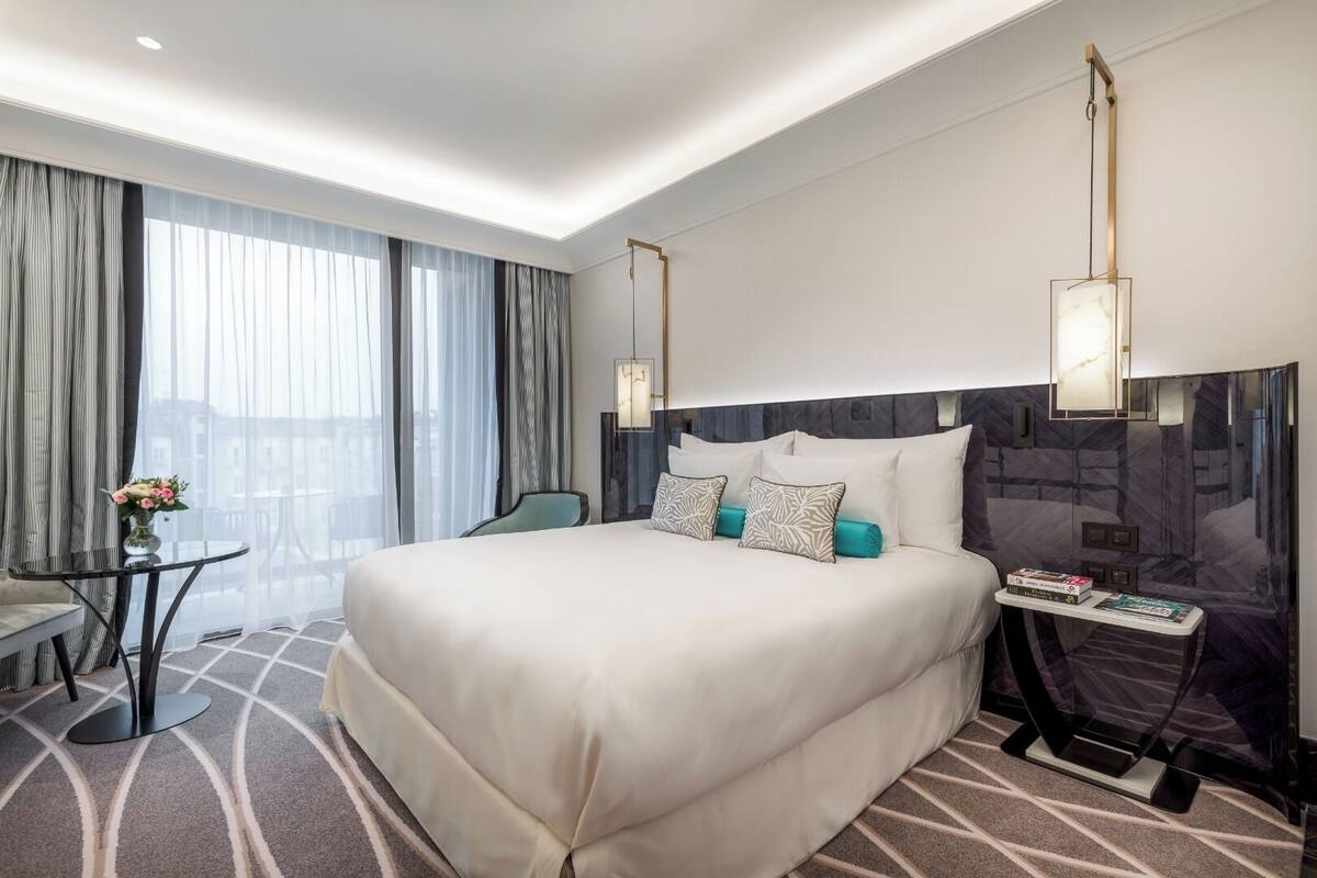 Un hotel moderno con una cama grande, una mesita con un florero, una silla y vistas a la ciudad a través de amplias ventanas.