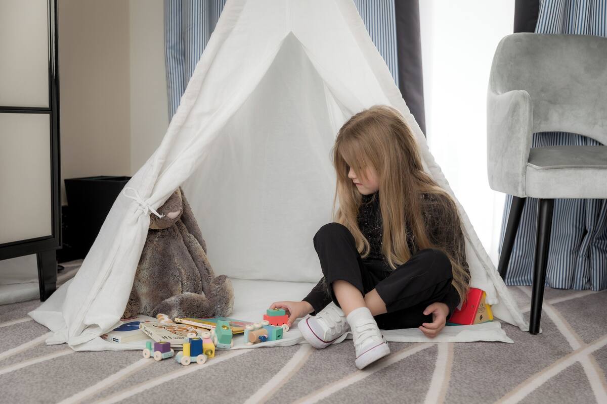 Un niño jugando con juguetes debajo de una tienda de campaña en una habitación con cortinas a rayas y una silla gris.