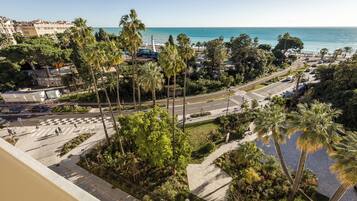 A coastal cityscape with a view of the sea, palm trees, and a road with vehicles.