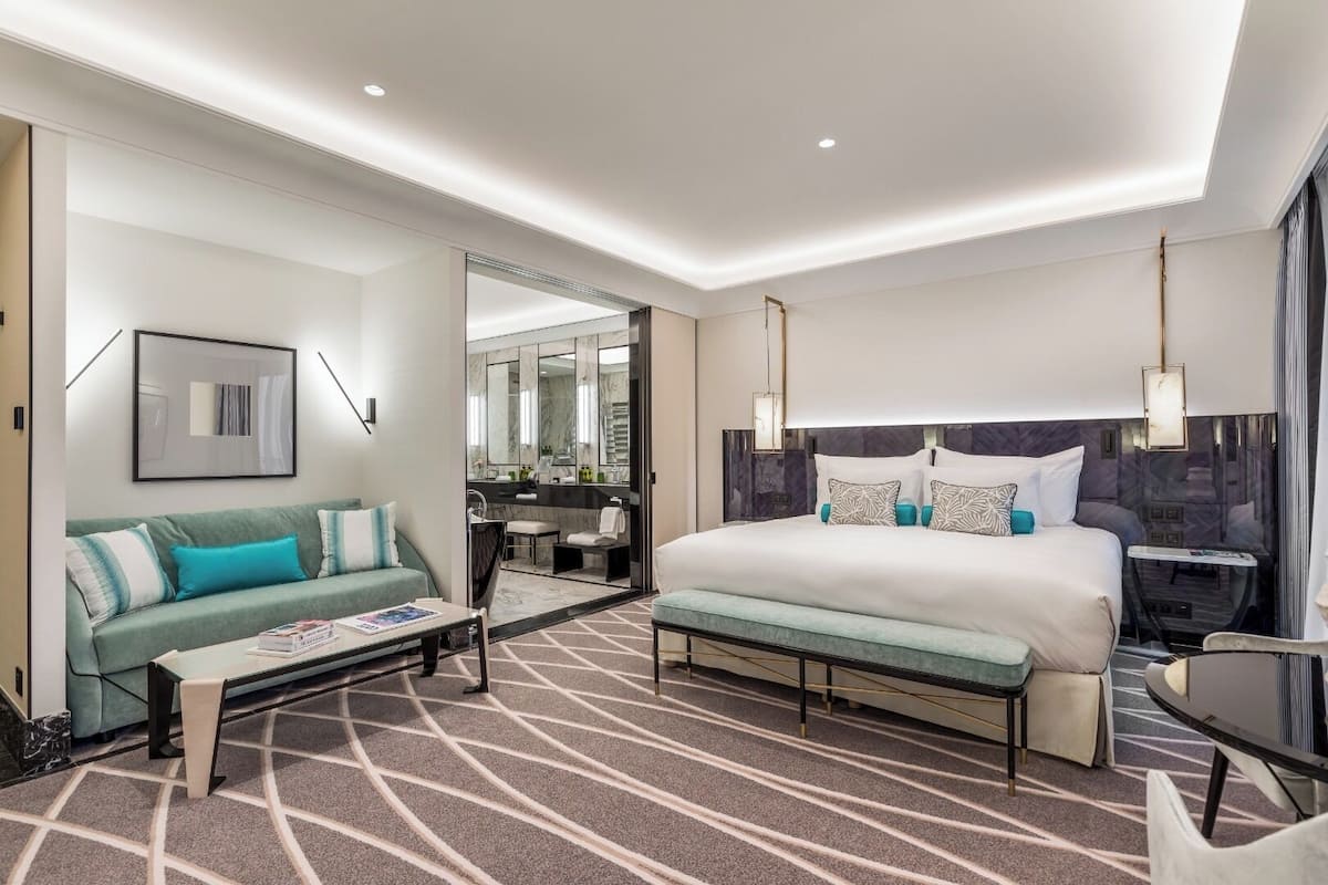 Habitación de hotel moderna con una cama grande, un sofá, una mesita y un baño visible a través de una puerta abierta.