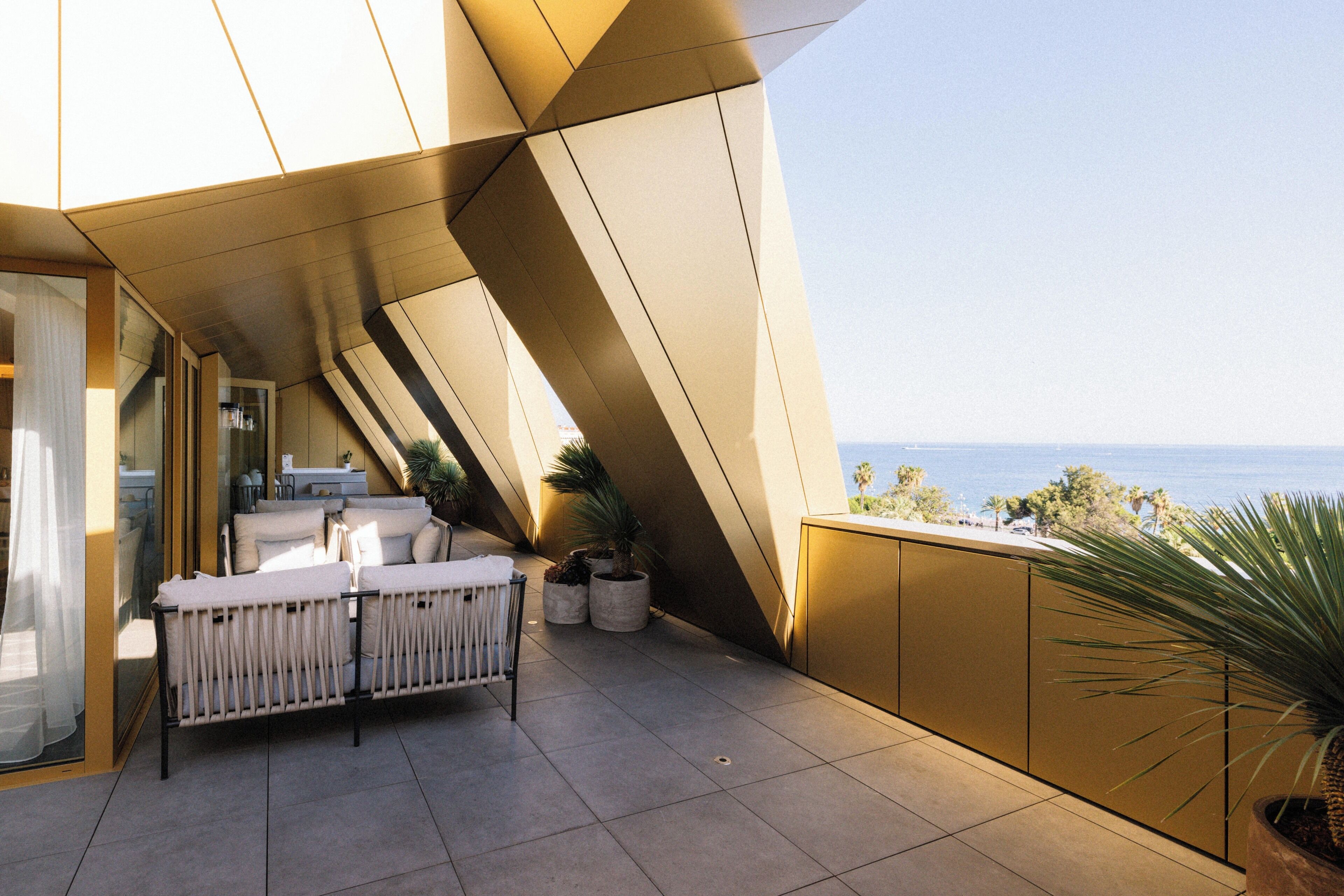 Een modern terras buiten met uitzicht op zee, met een bank en stoelen, plantenbakken en een geometrisch architectonisch ontwerp.