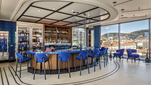 A modern bar with a curved ceiling, a large window offering a city view, and blue bar stools.