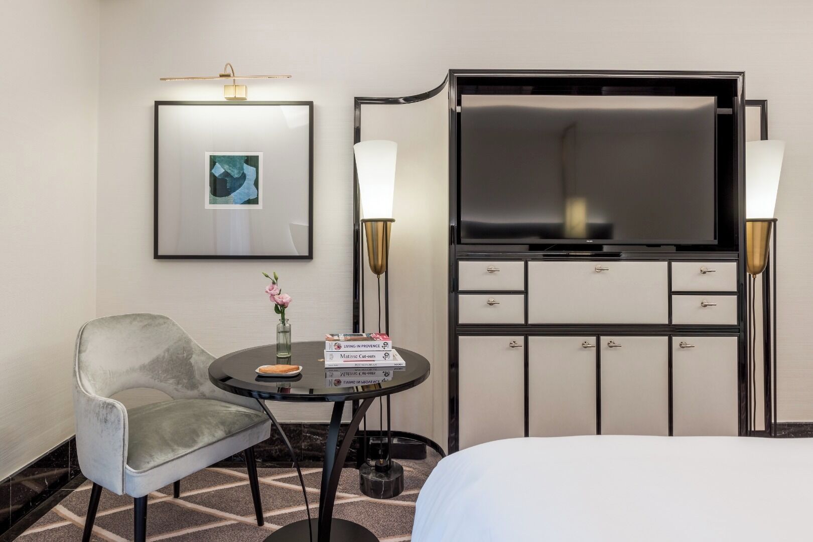 Habitación de hotel con cama, escritorio con silla, televisor, minibar y un cuadro en la pared.