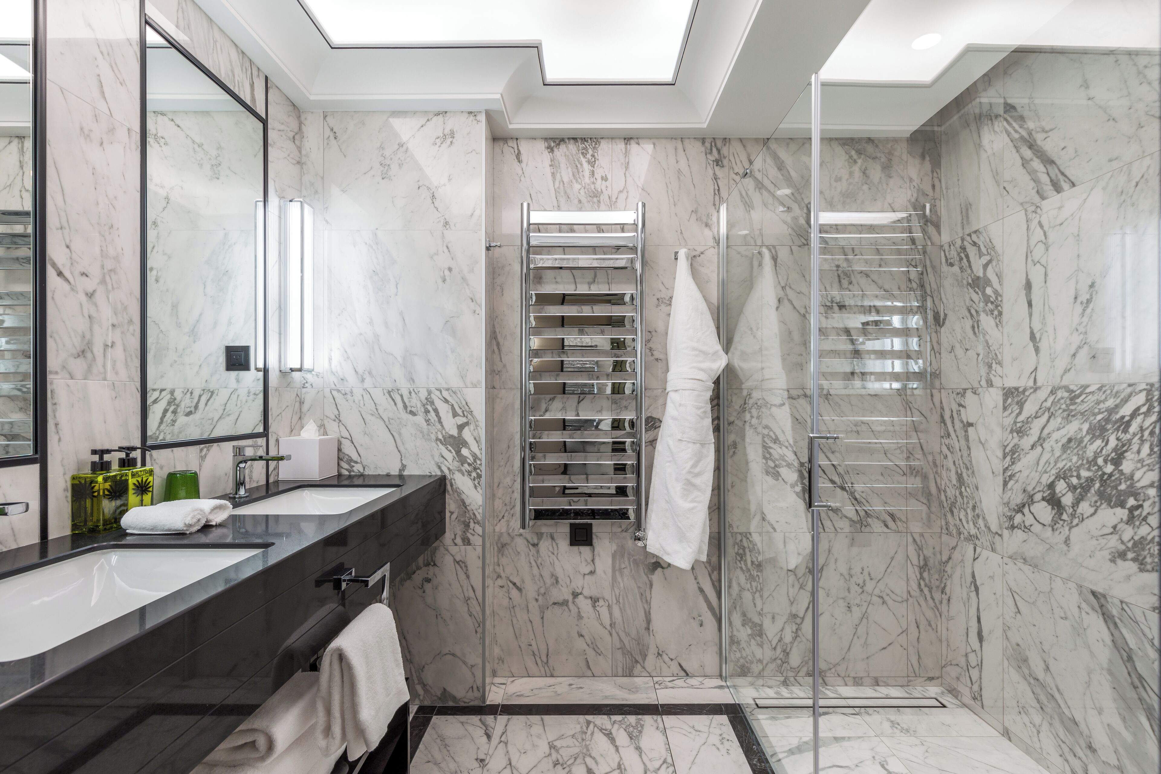 A modern bathroom with marble walls, a large mirror, dual sinks, a glass shower enclosure, and a towel rack.