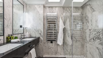 A modern bathroom with marble walls, a large mirror, dual sinks, a glass shower enclosure, and a towel rack.
