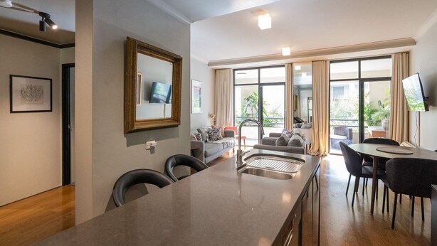 Eine moderne Küche mit Kochinsel, ein Essbereich mit Tisch und Stühlen, ein Wohnzimmer mit Sofa und Fernseher sowie große Fenster mit Vorhängen.
