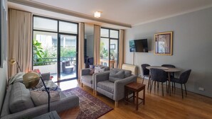Ein modernes Wohnzimmer mit einer Couch, Sesseln, einem Couchtisch und einem Essbereich mit Stühlen und einem Tisch. Ein Fernseher ist an der Wand befestigt und es gibt ein Fenster mit Vorhängen.