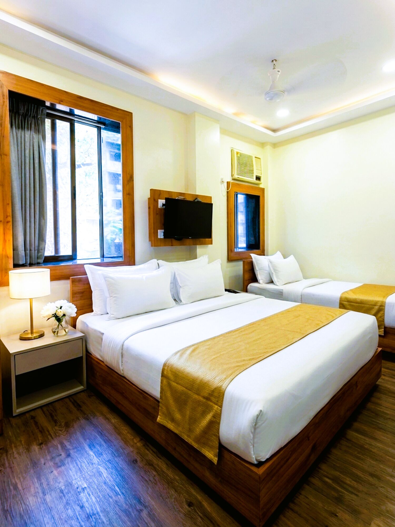 Una habitación de hotel con una cama grande, una cama más pequeña, un televisor, una ventana con cortinas, una mesita de noche con una lámpara y suelo de madera.