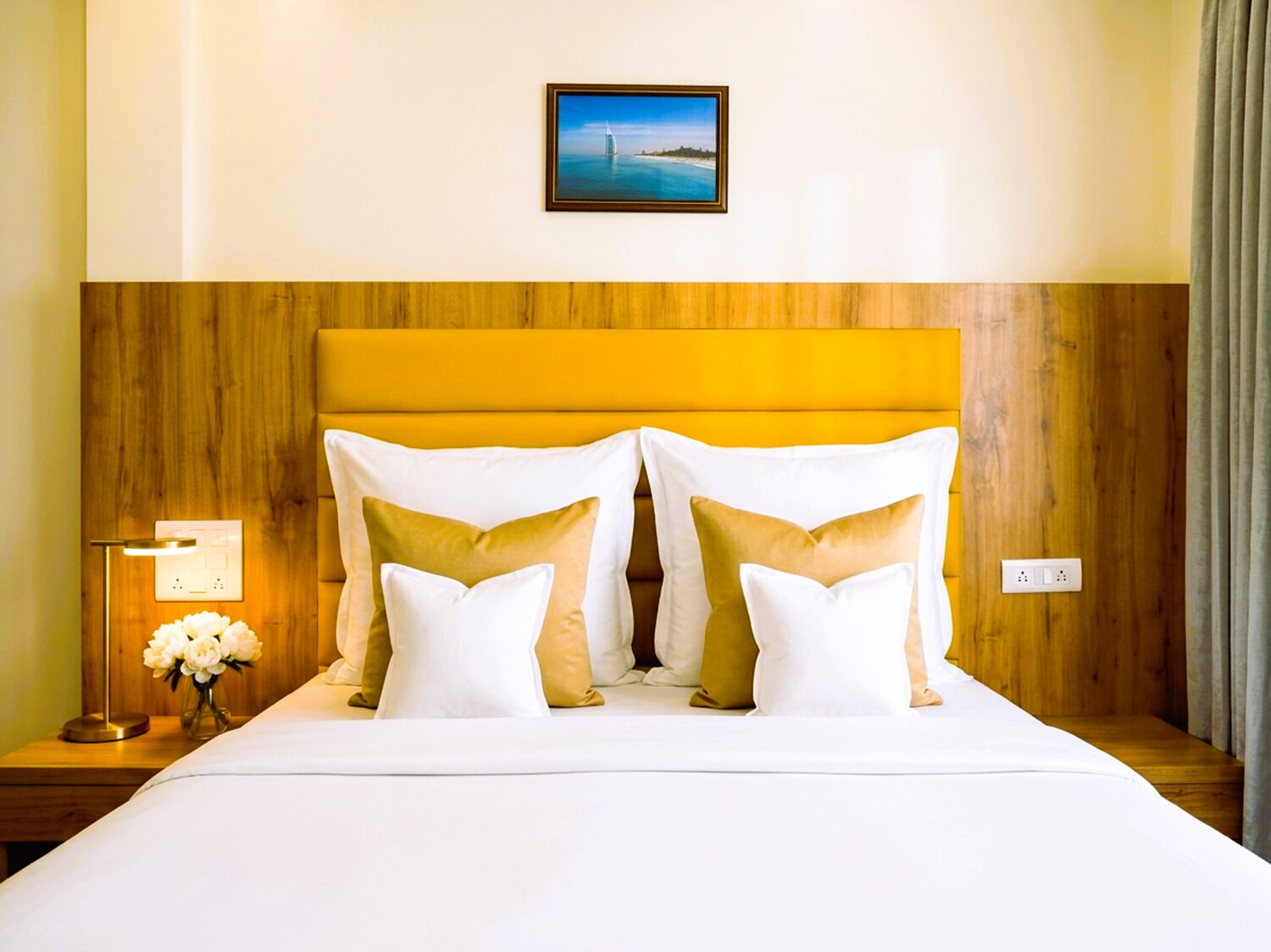 A hotel room with a wooden headboard, a bed with white and gold pillows, a nightstand with a lamp and flowers, and a framed picture on the wall.