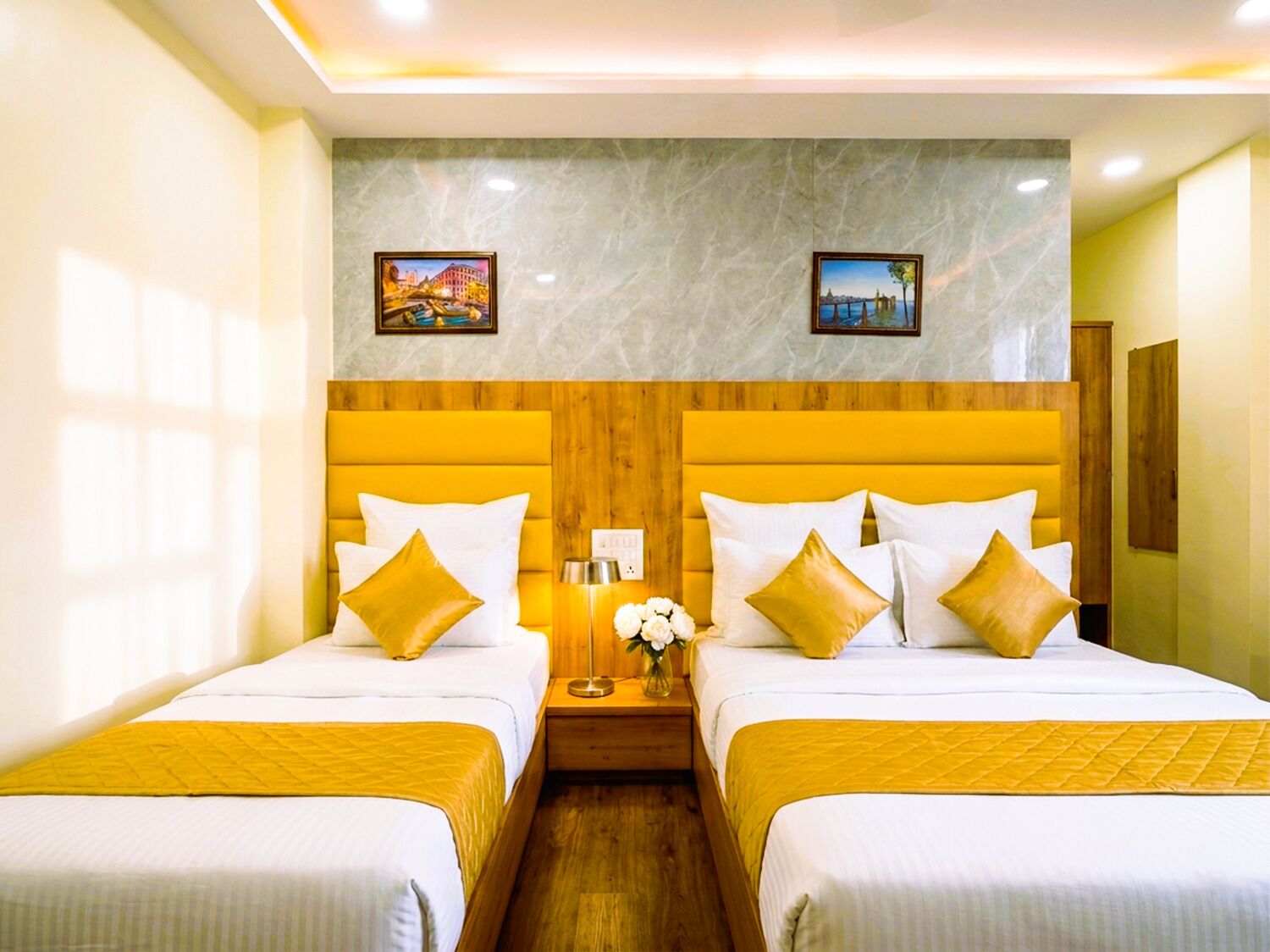 Habitación de hotel con dos camas, cabeceros de madera, ropa de cama blanca, una mesita de noche con lámpara y dos cuadros en la pared.