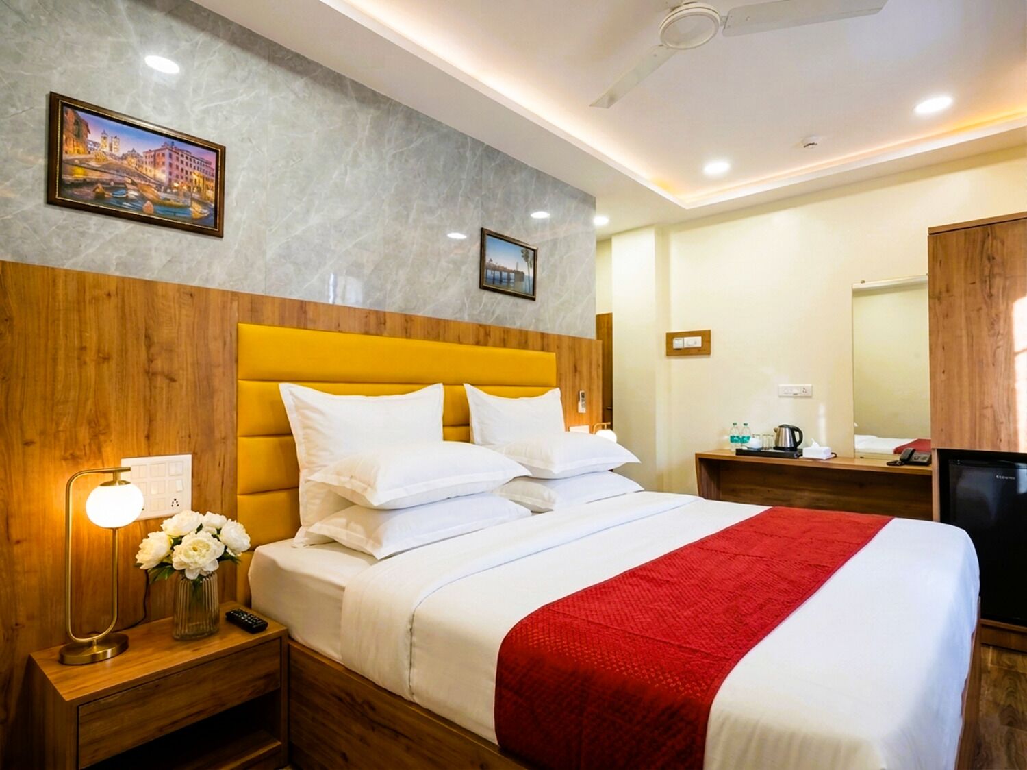 A hotel room with a bed, a nightstand with a lamp, a vase of flowers, a red throw blanket, and a television.