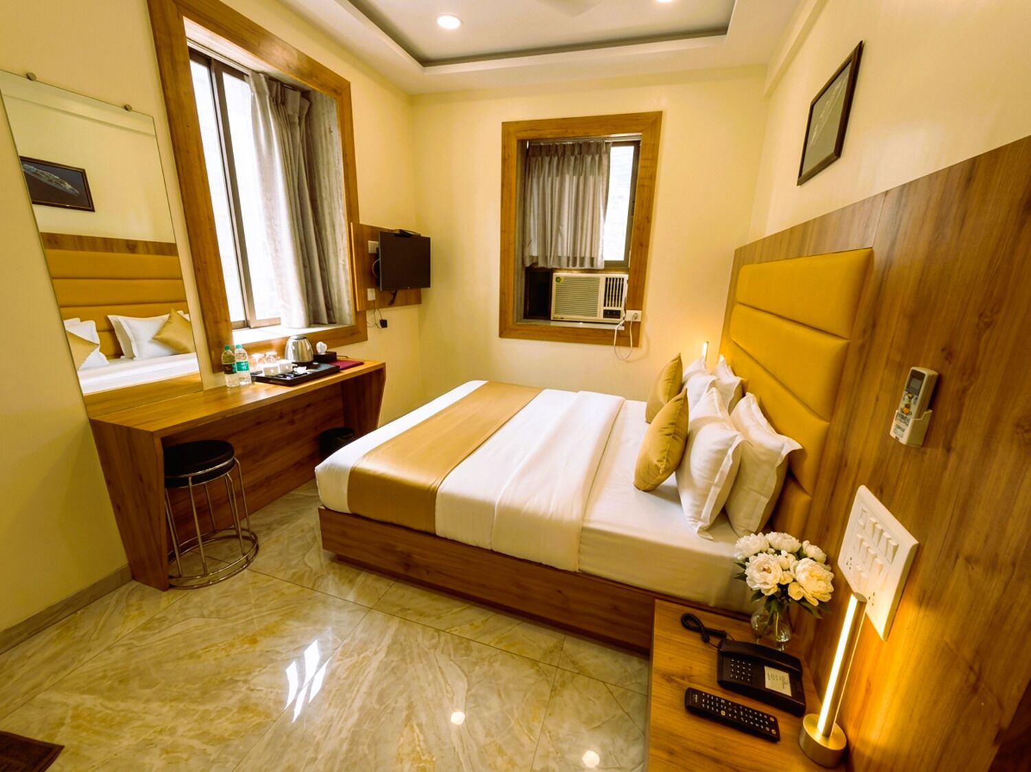 A hotel room with a bed, a desk with a chair, a mirror, a window with curtains, an air conditioning unit, and a bedside table with a vase of flowers.