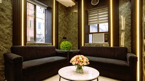 Un hall d’entrée moderne avec deux canapés sombres, une table ronde ornée d’un vase de fleurs et une grande fenêtre avec des stores.