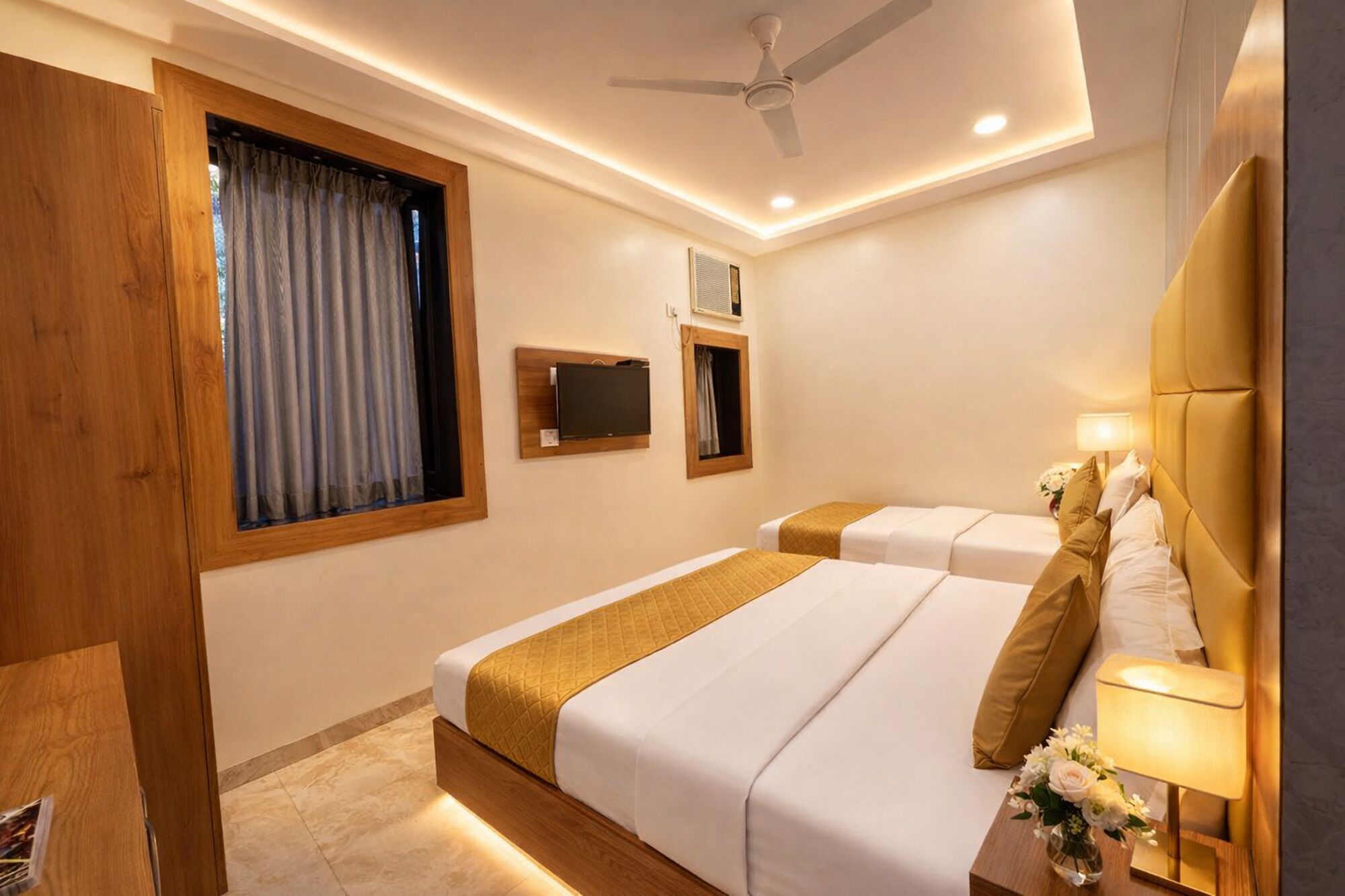 Une chambre d’hôtel avec deux lits, un ventilateur de plafond, des spots encastrés, une télévision et une table de chevet avec une lampe et des fleurs.