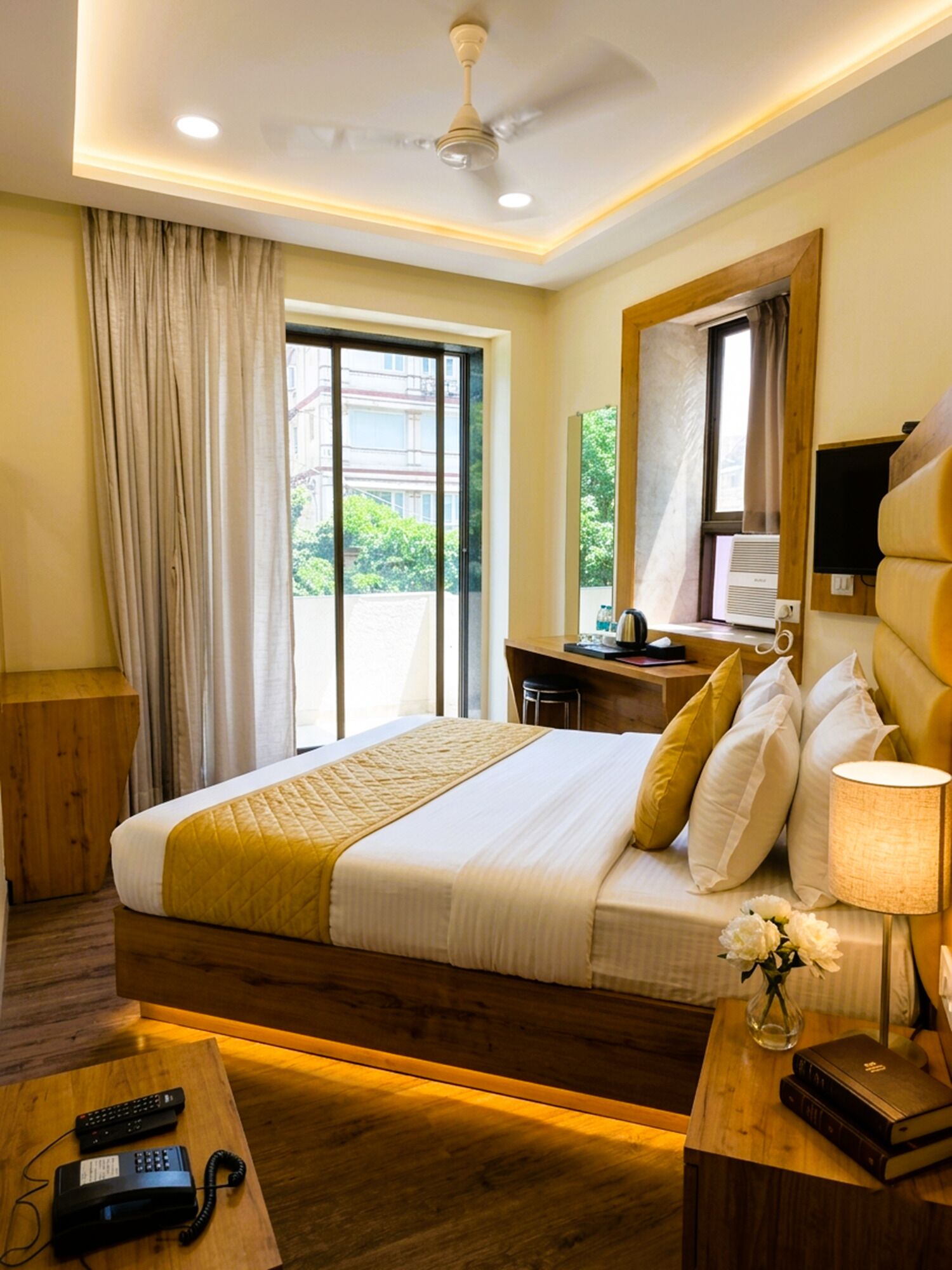 A hotel room with a bed, a desk, a chair, a TV, a lamp, a vase with flowers, and a window with curtains.