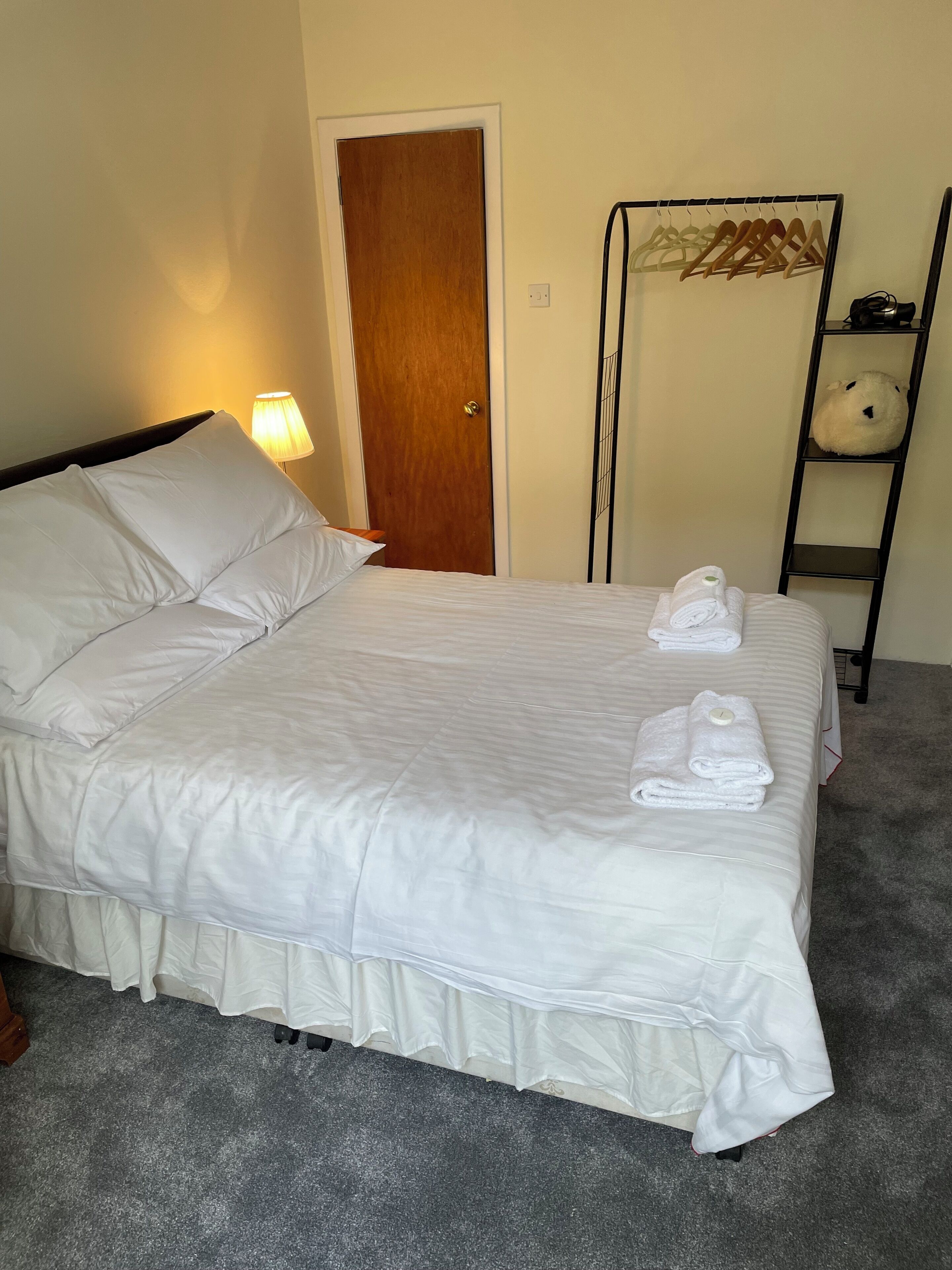 Una cama bien tendida con ropa de cama y toallas blancas, una puerta de madera, una lámpara y un estante con un peluche.
