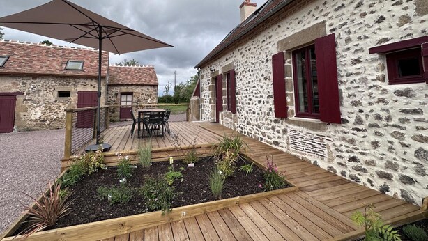 A stone building with red shutters, a wooden deck with a table and chairs, and a garden bed with various plants.