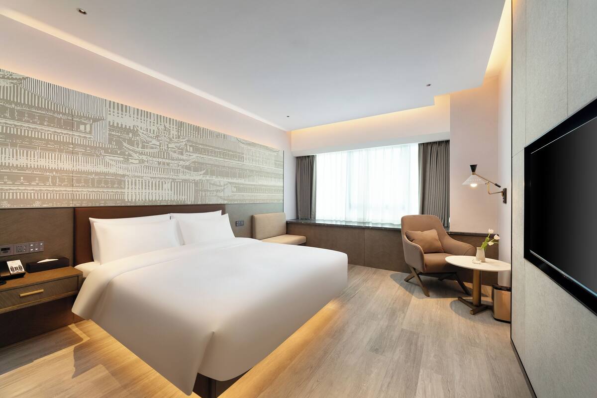 Una camera d'albergo moderna con un letto grande, una zona salotto con una poltrona e un tavolino, una TV a schermo piatto e un'opera d'arte appesa al muro.