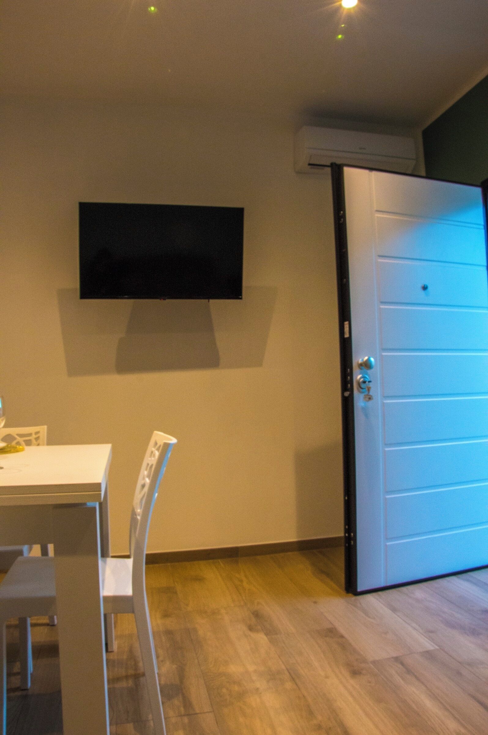 Ein Zimmer mit weißer Tür, einem an der Wand befestigten Flachbildfernseher, einem weißen Tisch mit Stuhl und Holzboden.