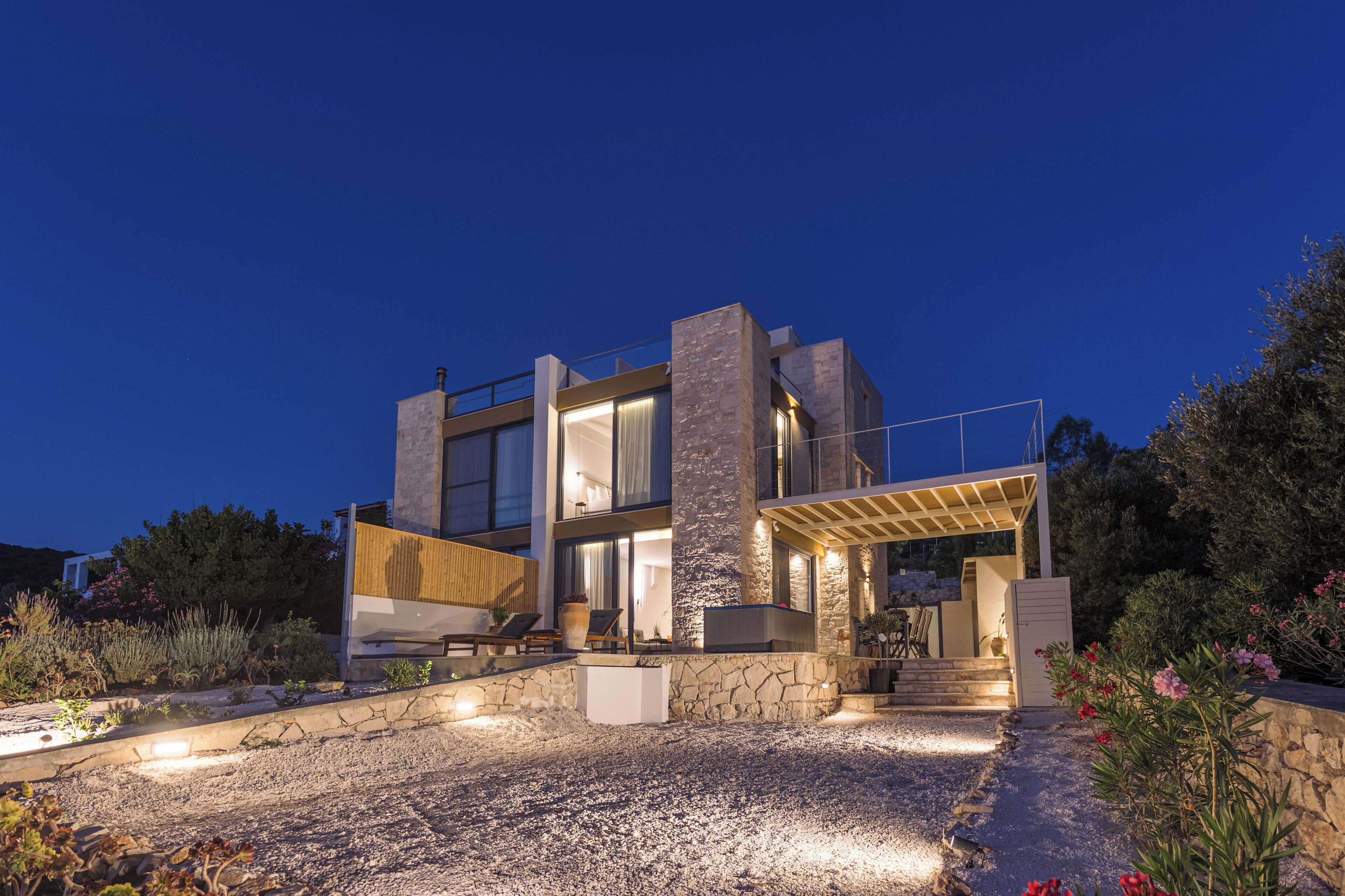A modern house with a stone facade, large windows, and a well-lit outdoor area with seating and plants.