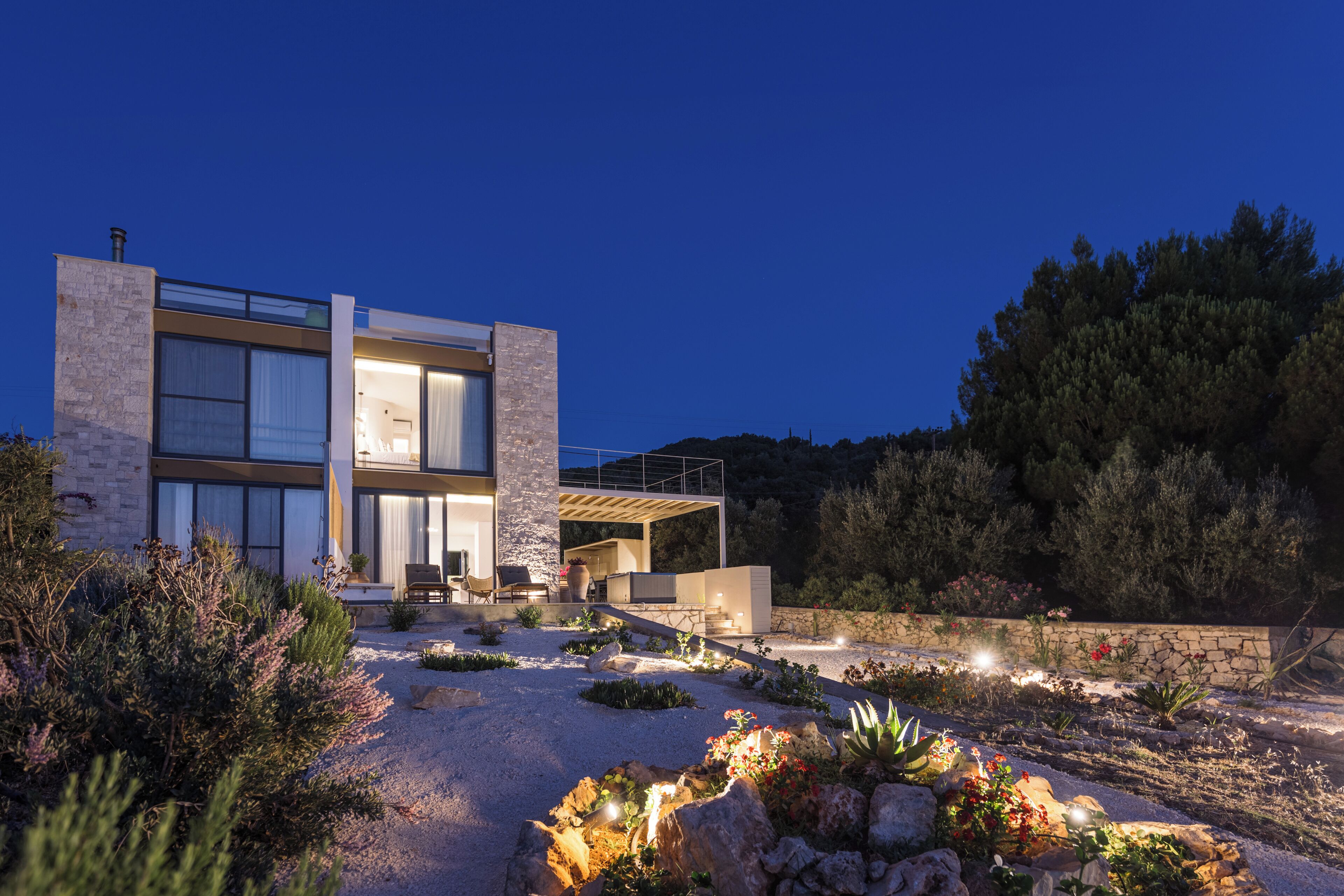 A modern house with a large glass facade, a stone exterior, and a landscaped garden with various plants and rocks.