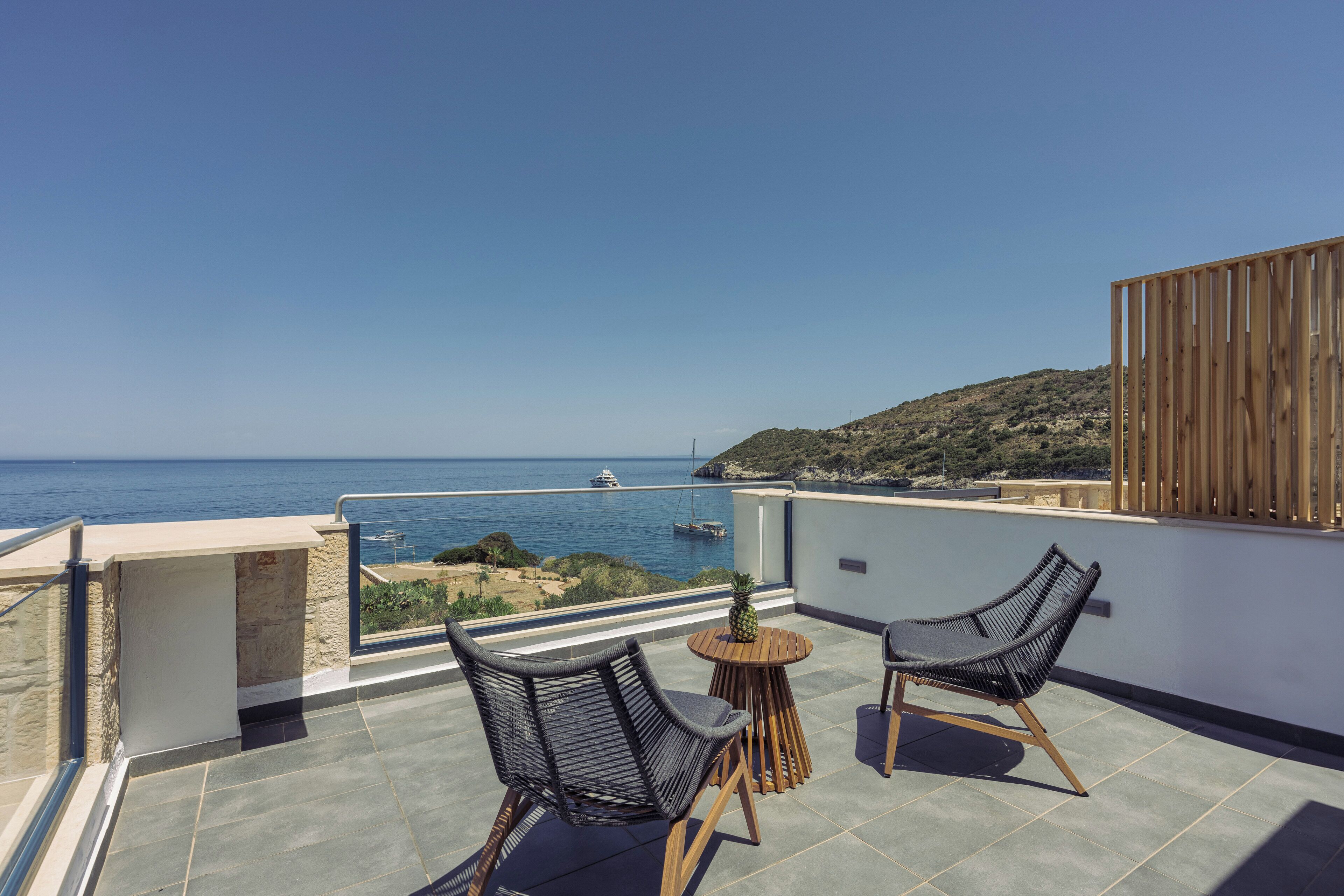 A balcony with two wicker chairs, a small round table, and a view of the sea and a boat in the distance.