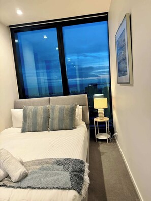 A hotel room with a large window, a bed with pillows, a bedside table with a lamp, and a framed picture on the wall.