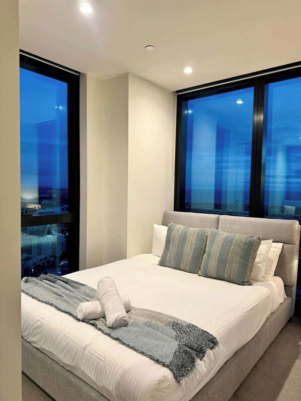 A modern hotel room with a large bed, a view of the cityscape through floor-to-ceiling windows, and a city skyline visible through the window.