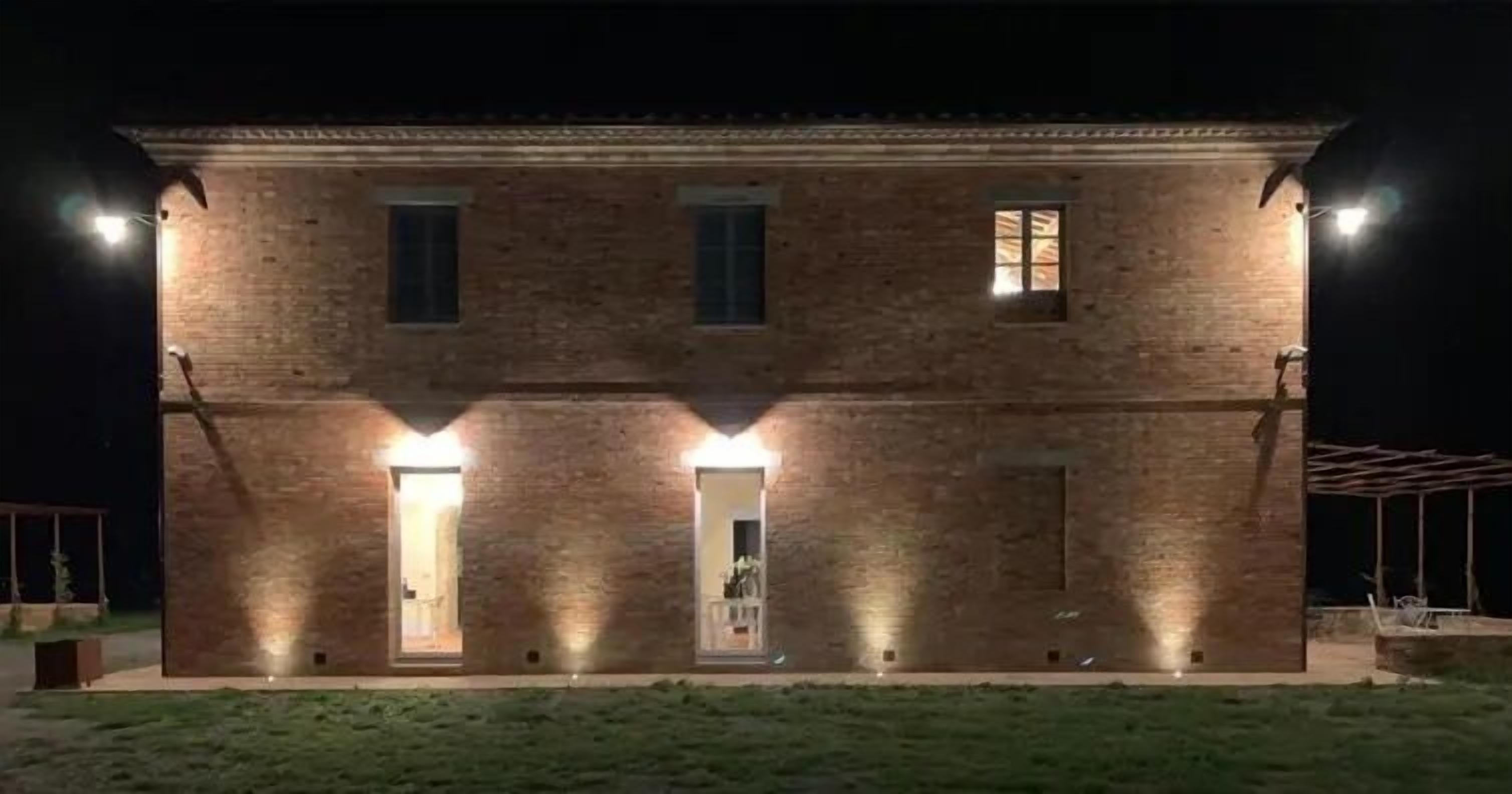 A two-story brick building with illuminated windows and doors, a flat roof, and a paved front area.
