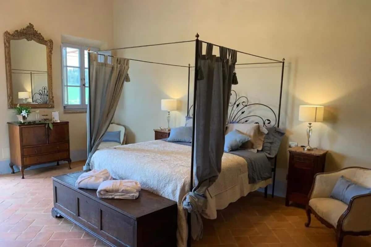 A four-poster bed with a canopy, a wooden dresser, a chair, and a nightstand with lamps.