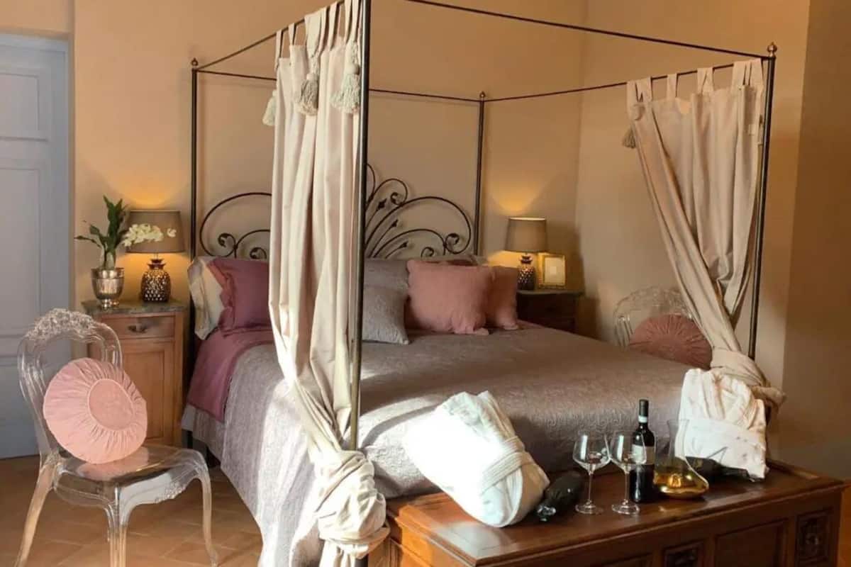 A four-poster canopy bed with a wooden headboard, two bedside tables with lamps, a chair with a cushion, and a wooden nightstand with a vase and a bottle.