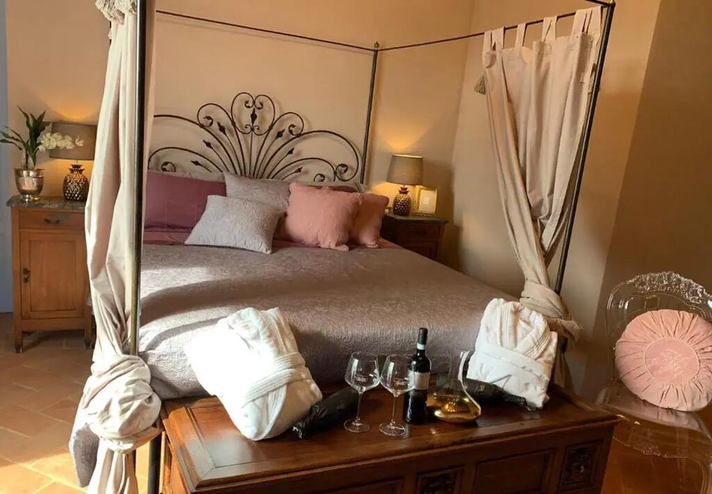 A four-poster bed with a canopy, a bedside table with wine glasses and a bottle, and a nightstand with a lamp and decorative items.
