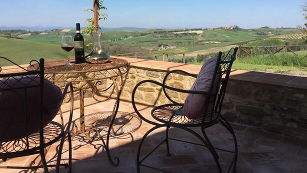 A patio with a table and chairs, a bottle of wine, and a glass of red wine. The patio overlooks a scenic landscape with rolling hills and a distant building. - Cordella in Valdorcia Truffle & Olive Oil Resort - Podere Borgasso (Montalcino)