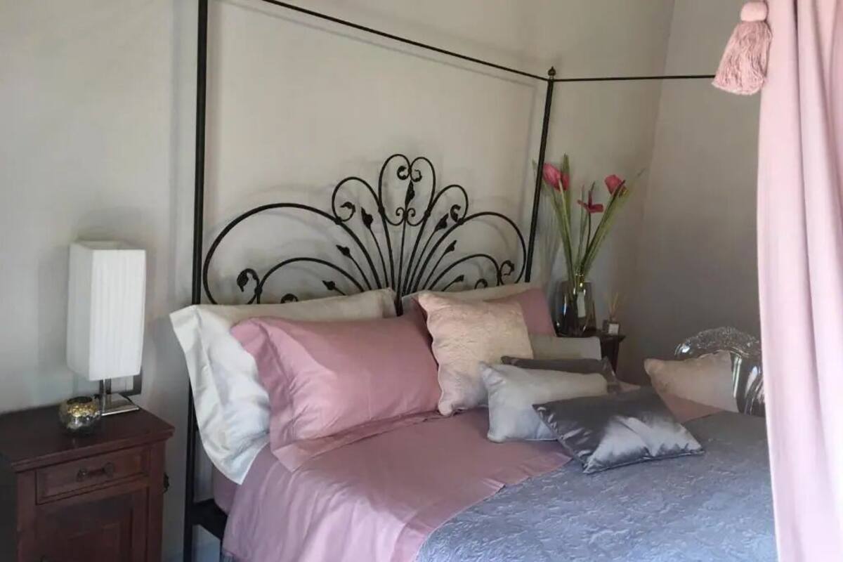 A bedroom with a metal headboard, a bed with pink and grey bedding, a bedside table with a lamp, and a vase with flowers.